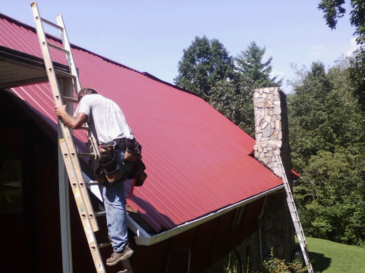 Roofer on a ladder beside a red metal roof and stone chimney — Soffit & Fascia Repair by Roofing Brothers in Suffield